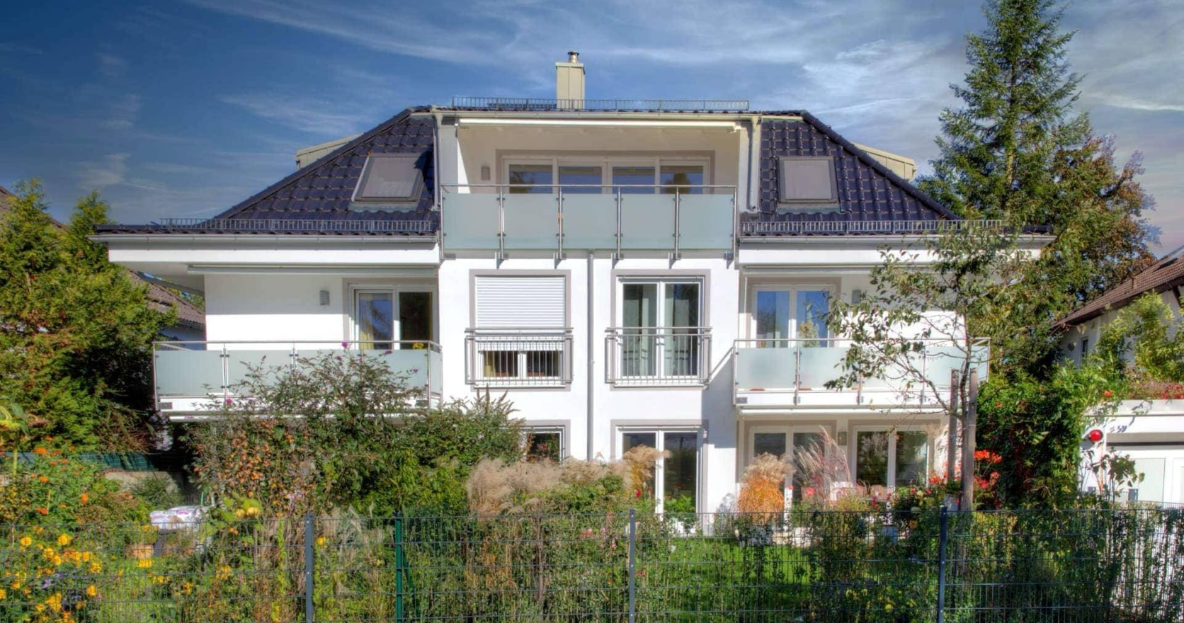 Weißes Einfamilienhaus mit dunklem Walmdach, Balkon und blühendem Garten, Außenansicht einer Münchner Immobilie