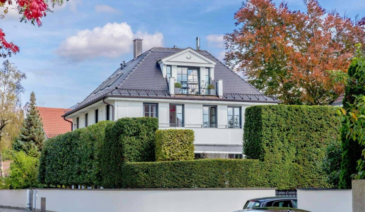 Weiße Villa mit dunklem Dach hinter hoher grüner Hecke und buntem Herbstlaub in München