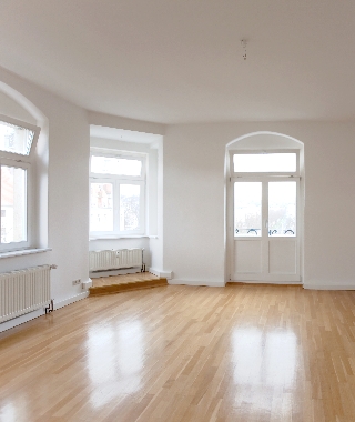 Leerer, heller Altbauraum mit glänzendem Parkettboden, Rundbogenfenstern und Stuckdecke.
