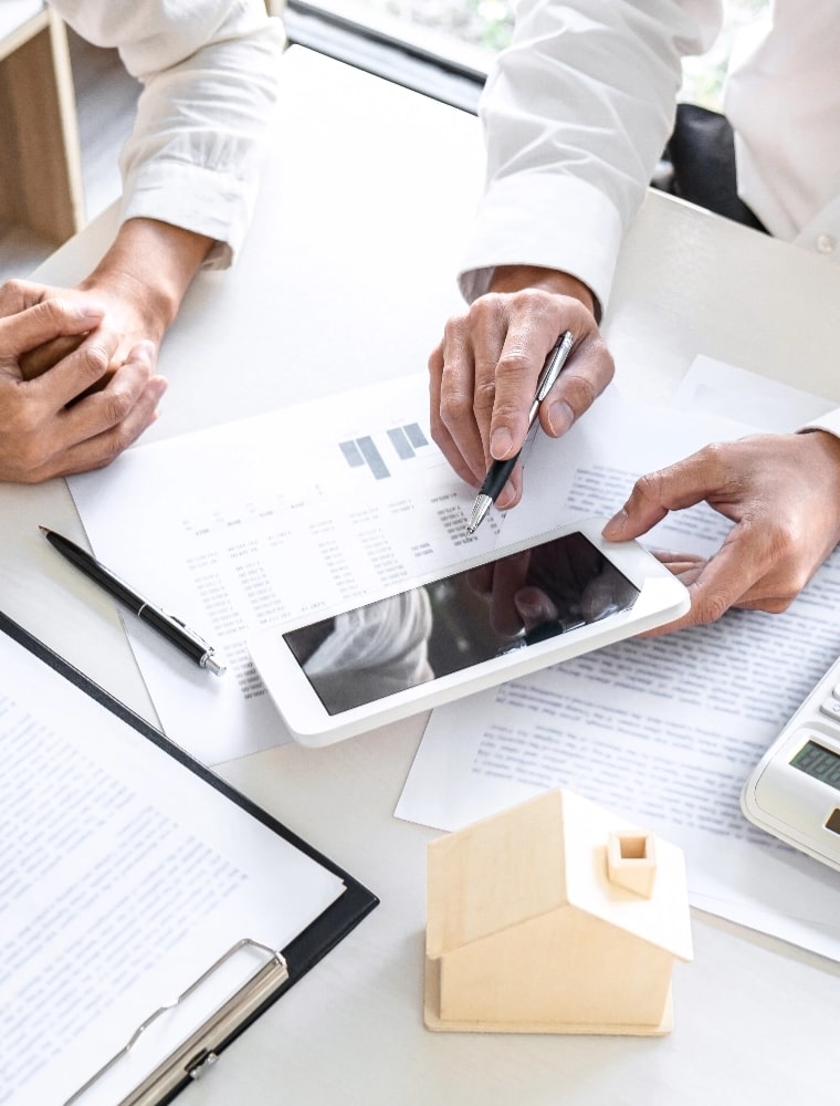 Two people are discussing a diagram on a tablet, with a small wooden house model and paperwork in the foreground.