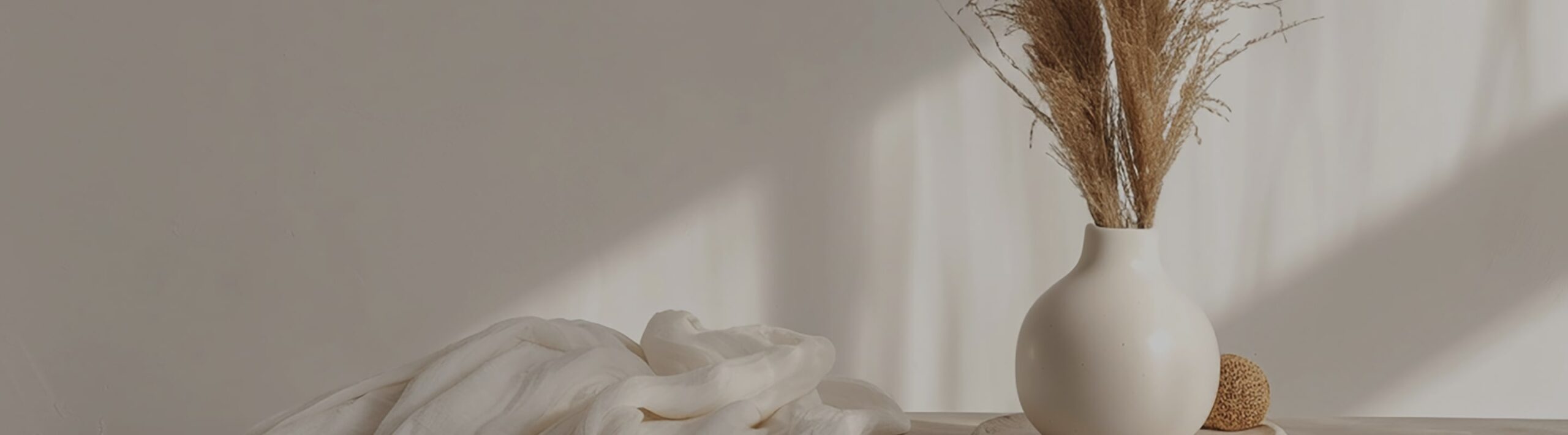 Bright, calm living scene with a white vase, dried grasses, and soft linen fabric in warm light.