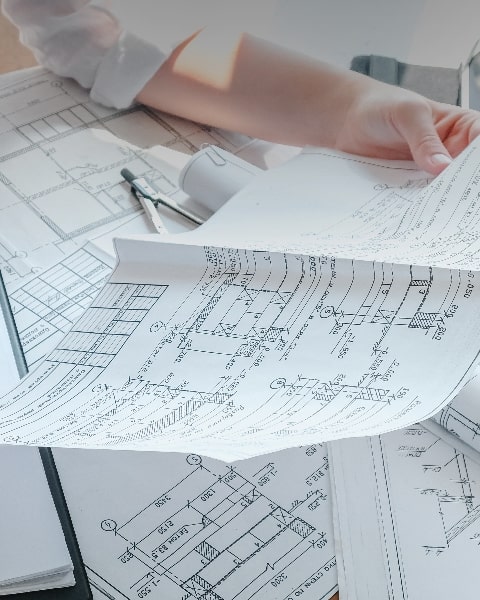 Eine Hand blättert in ausgebreiteten technischen Bauplänen mit Grundrisszeichnungen und Notizen.