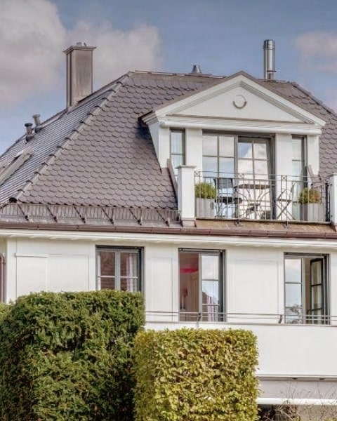 Weiße Villa mit Dachterrasse, Säulen-Erker und gepflegter Heckenbepflanzung im Vordergrund.