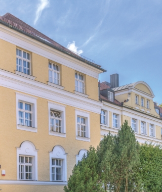 Klassizistische gelbe Altbaufassade mit mehreren Fensterreihen und grünen Bäumen im Vordergrund.