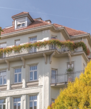 Historische Fassade mit begrüntem Balkongeländer, Fensterläden und rotem Ziegeldach unter blauem Himmel.