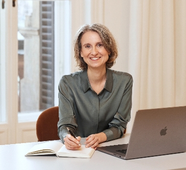 Katerina Rogers sitzt lächelnd am weißen Schreibtisch, schreibt Notizen neben ihrem aufgeklappten Laptop.