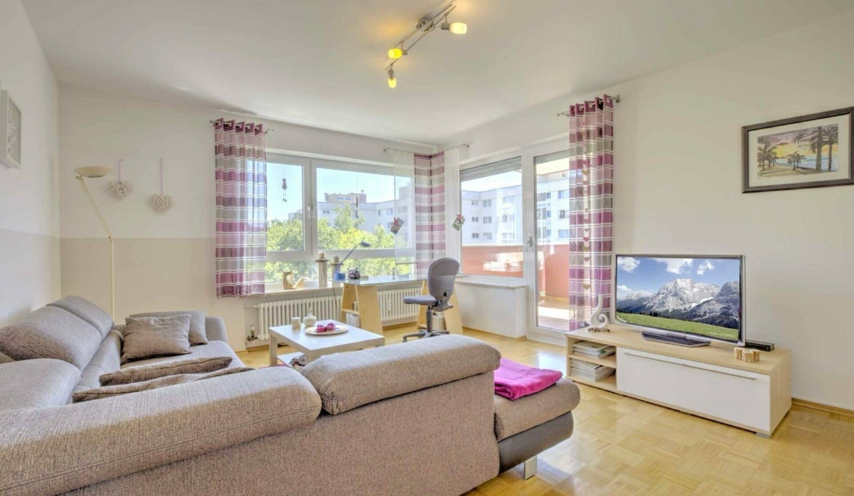 Helles Wohnzimmer mit beigem Sofa, Fernseher und großem Fenster mit Ausblick in einer Münchner Wohnung