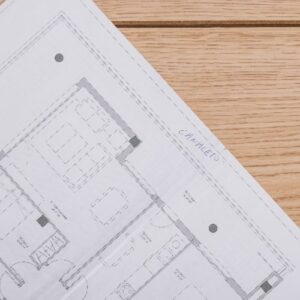 Ausschnitt eines Bauplans mit Grundrisslinien, der auf einem hellen Holzdielenboden ausgebreitet liegt.