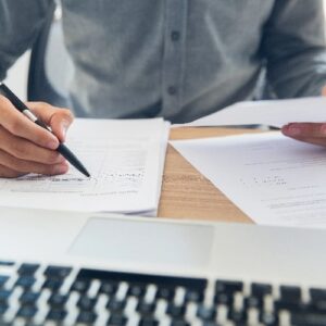 Ein Geschäftsmann sitzt am Schreibtisch und unterschreibt mit einem Kugelschreiber Vertragsunterlagen neben einem Laptop.