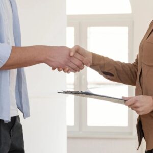 Two people are shaking hands in a bright, empty apartment, one holding a clipboard with documents.