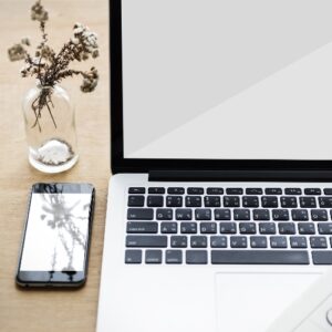 Laptop mit leerem Bildschirm neben Smartphone auf Holztisch, daneben ein Glas mit getrockneten Blumen.