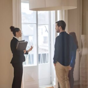 Eine Immobilienmaklerin im schwarzen Blazer erklärt einem jungen Paar in einer hellen Wohnung die Räumlichkeiten.