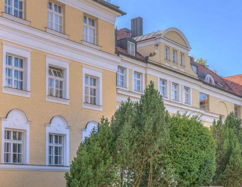 Gelbe klassizistische Fassade mit Dreiecksgiebel und Nadelbäumen im Vorgarten