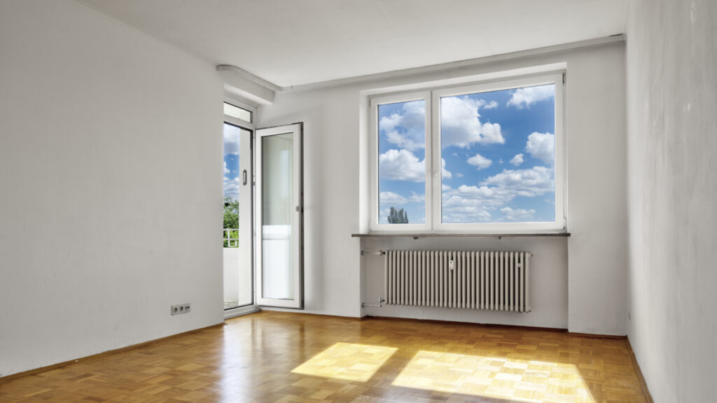 Leeres, helles Zimmer mit Balkontür, Heizkörper und Parkettboden ohne Möblierung