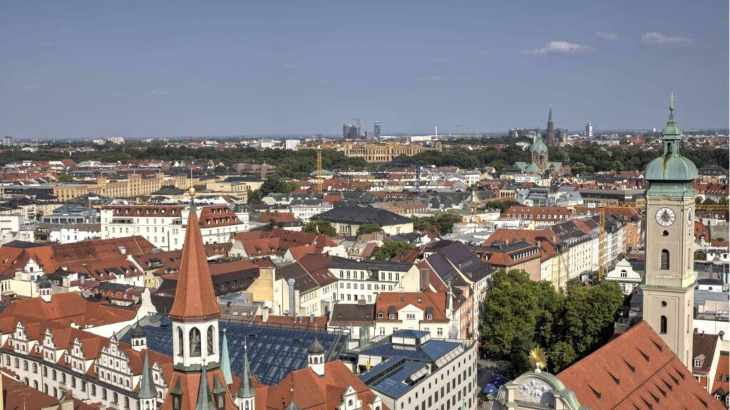 Blick über die Münchner Dächer mit grünem Kirchturm im Vordergrund und Stadtsilhouette im Hintergrund