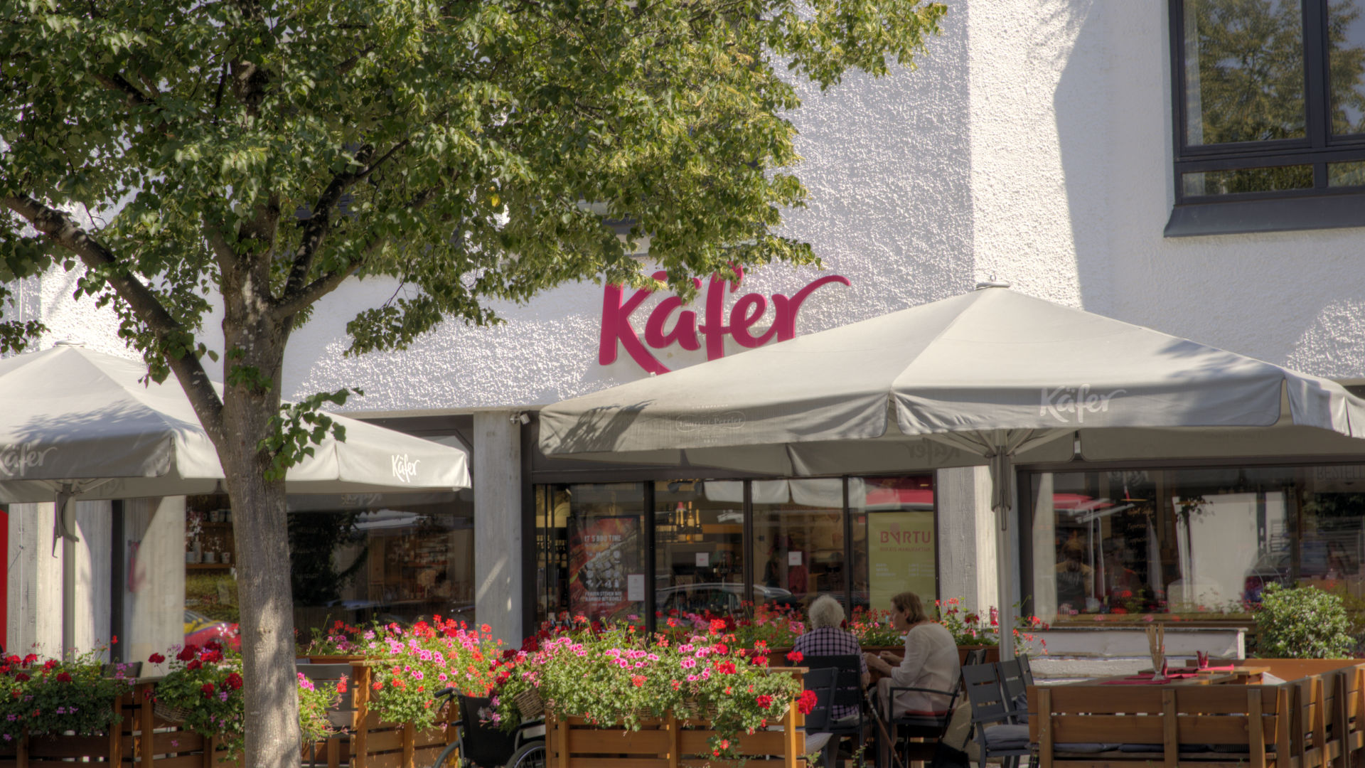 Eingang der Feinkost-Käfer-Filiale mit Sonnenschirmen und Sitzgelegenheiten im Münchner Stadtteil Solln
