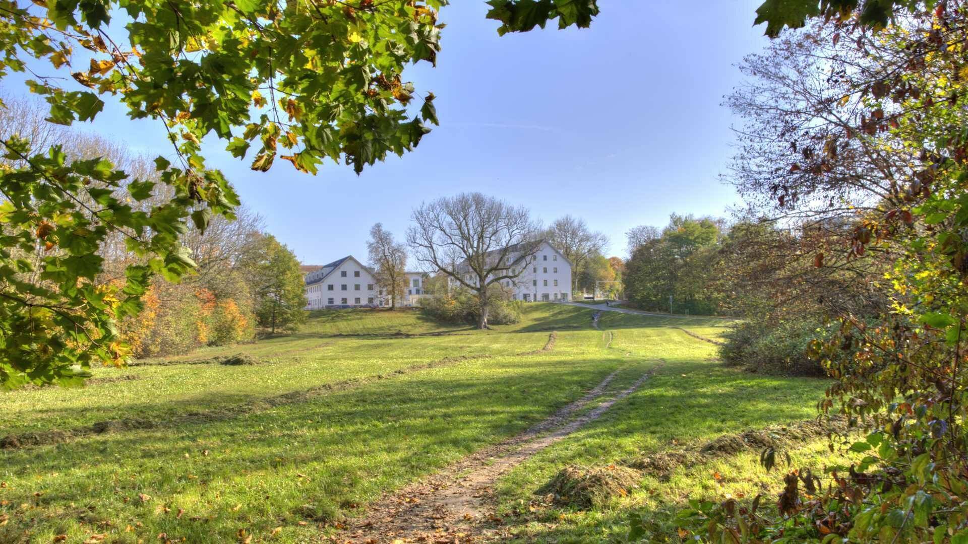 Grüne Wiese mit historischem Gebäude im Hintergrund, eingerahmt von herbstlichen Bäumen