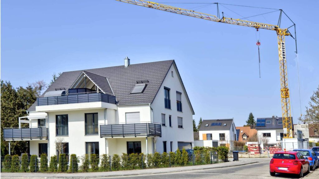 Neu gebautes weißes Reihenhaus mit Baukran im Hintergrund und rotem Auto davor