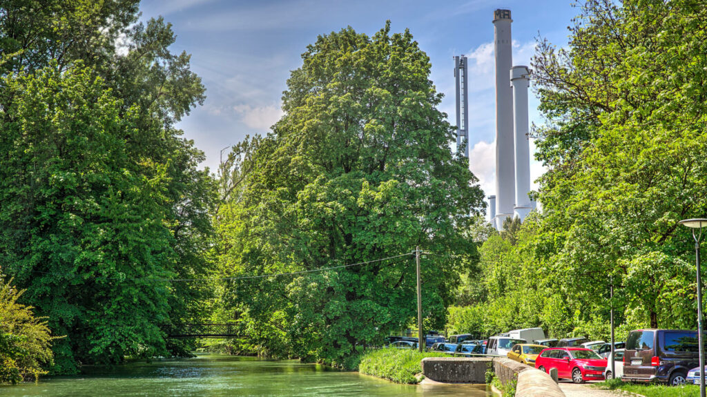 Grüner Flusskanal mit Bäumen, im Hintergrund die Schornsteine eines Heizkraftwerks in München