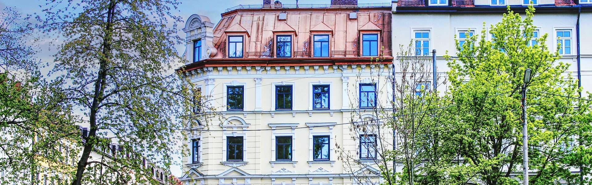 Weiße Altbaufassade mit rotem Ziegeldach zwischen Bäumen im Münchner Stadtteil Isarvorstadt