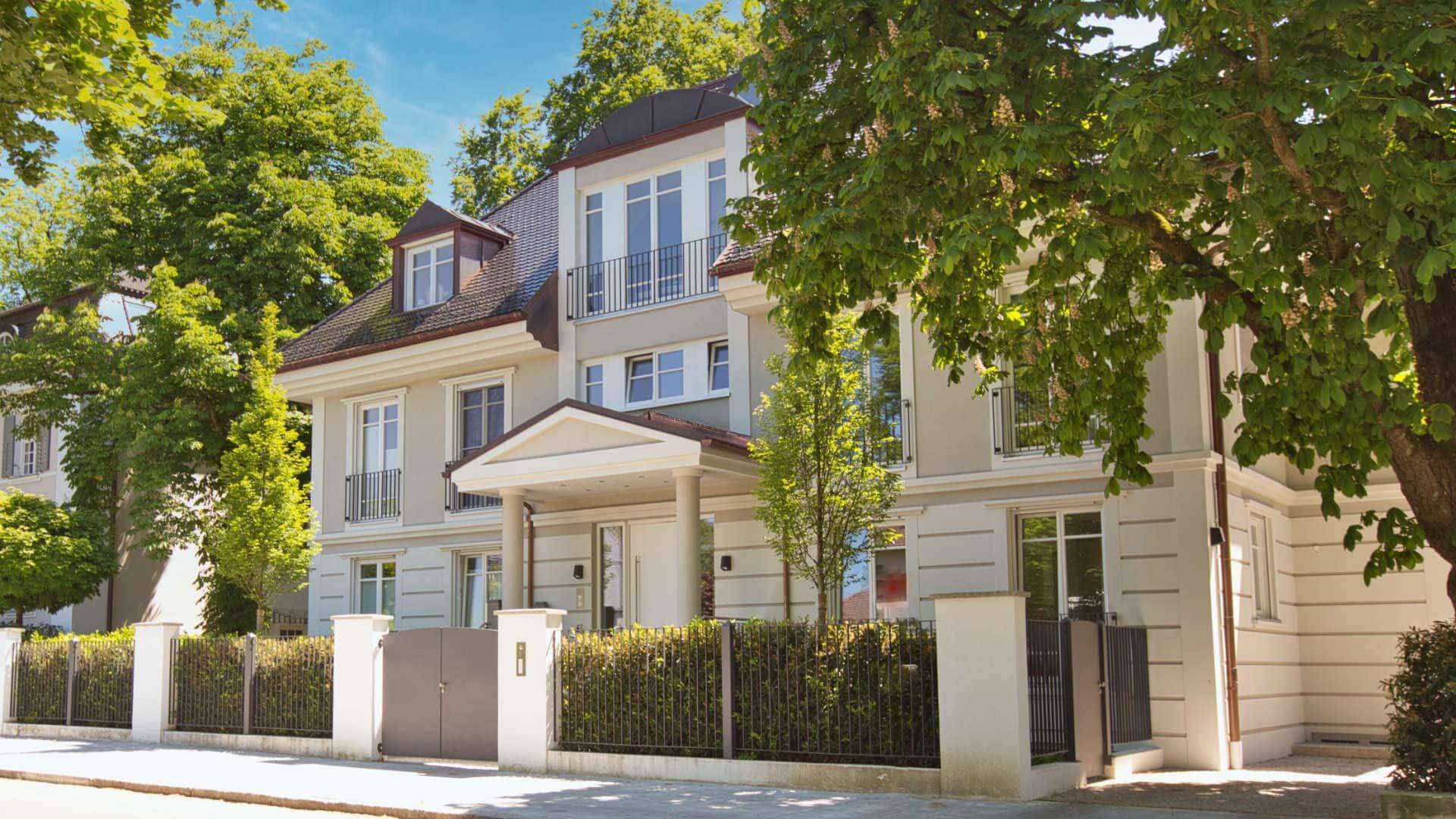 Cremefarbene Villa mit Mansarddach hinter schmiedeeisernem Zaun und Bäumen in München-Nymphenburg