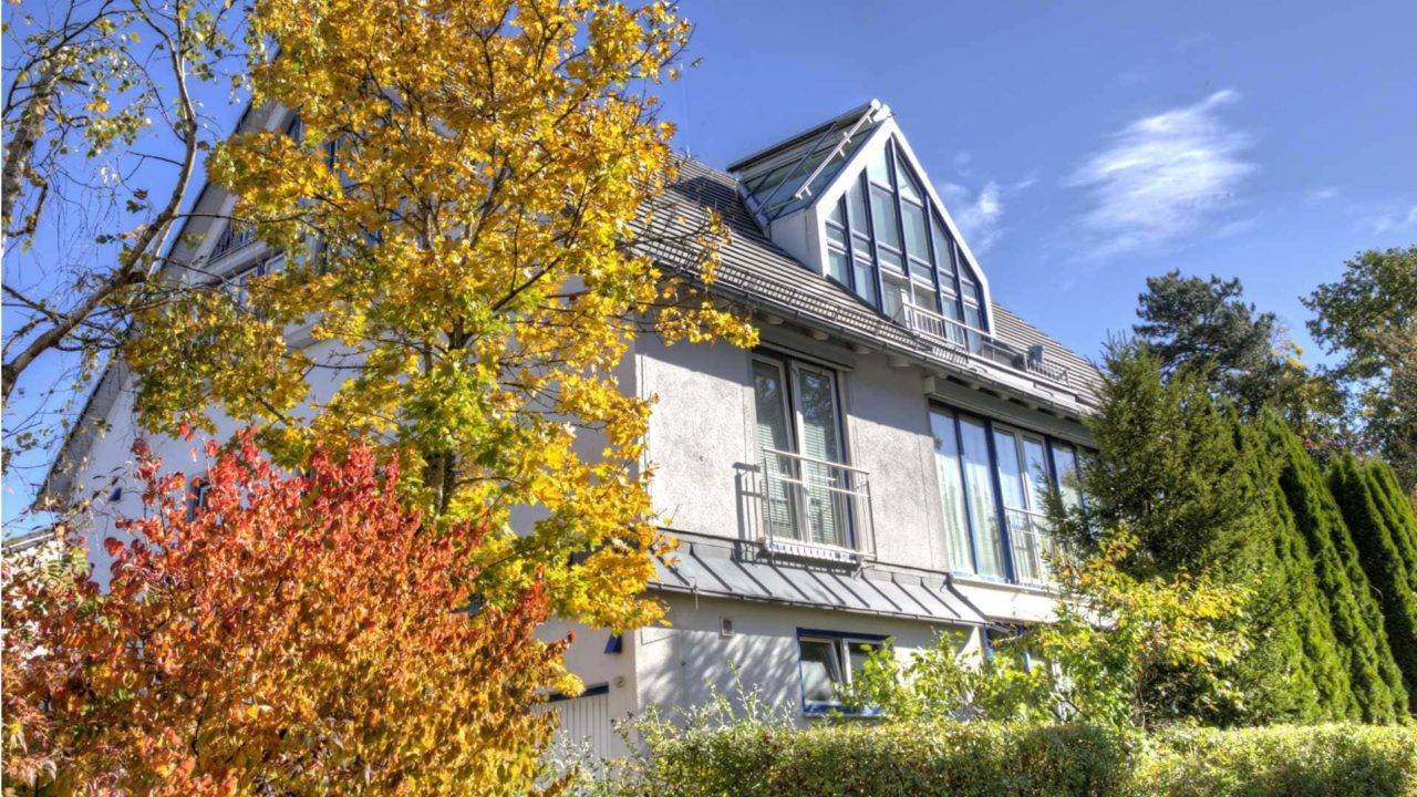 Weiße Villa mit spitzem Glasgiebel hinter buntem Herbstlaub – Symbolbild zu Immobilienpreisen in München-Trudering