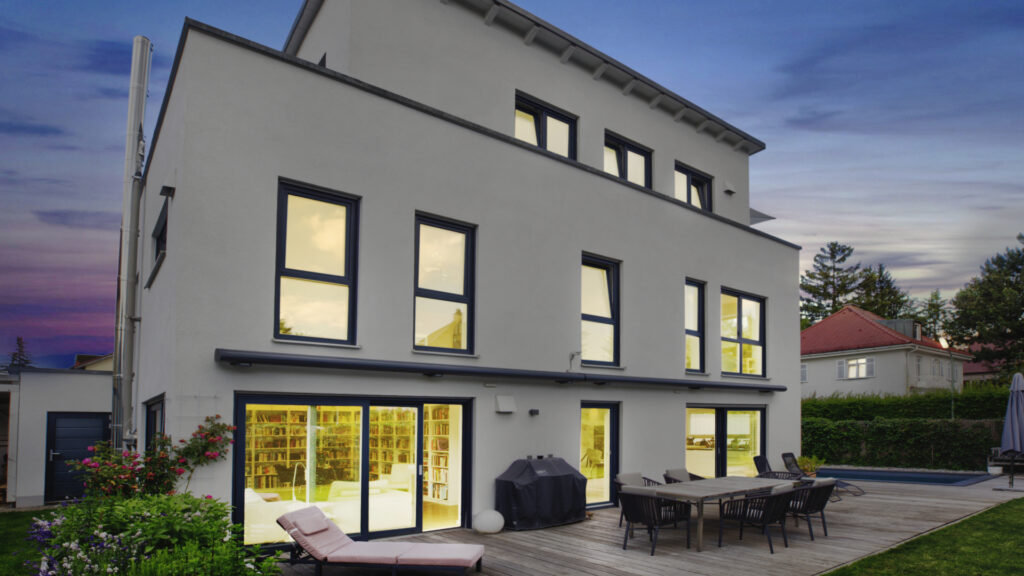 Modernes graues Neubauhaus mit beleuchteten Fenstern und Terrasse in der Abenddämmerung