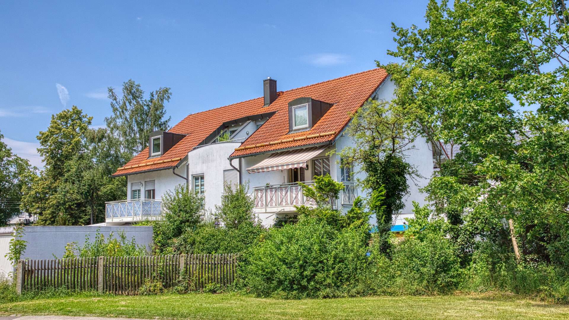 Weißes Haus mit rotem Ziegeldach und Balkon hinter Bäumen im Münchner Stadtteil Feldmoching