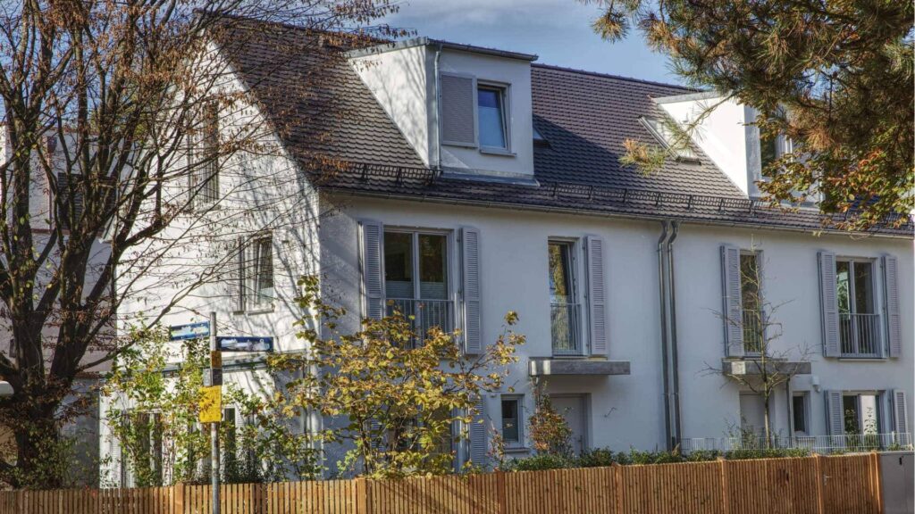 Weißes Wohnhaus mit grauen Fensterläden und Holzzaun im Herbstlaub – Symbolbild zu Betriebskosten als zweiter Miete