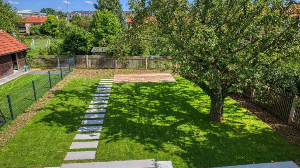 Großzügige Rasenfläche mit altem Baum, Steinweg und eingezäuntem Grundstück hinter einer Doppelhaushälfte in Bogenhausen