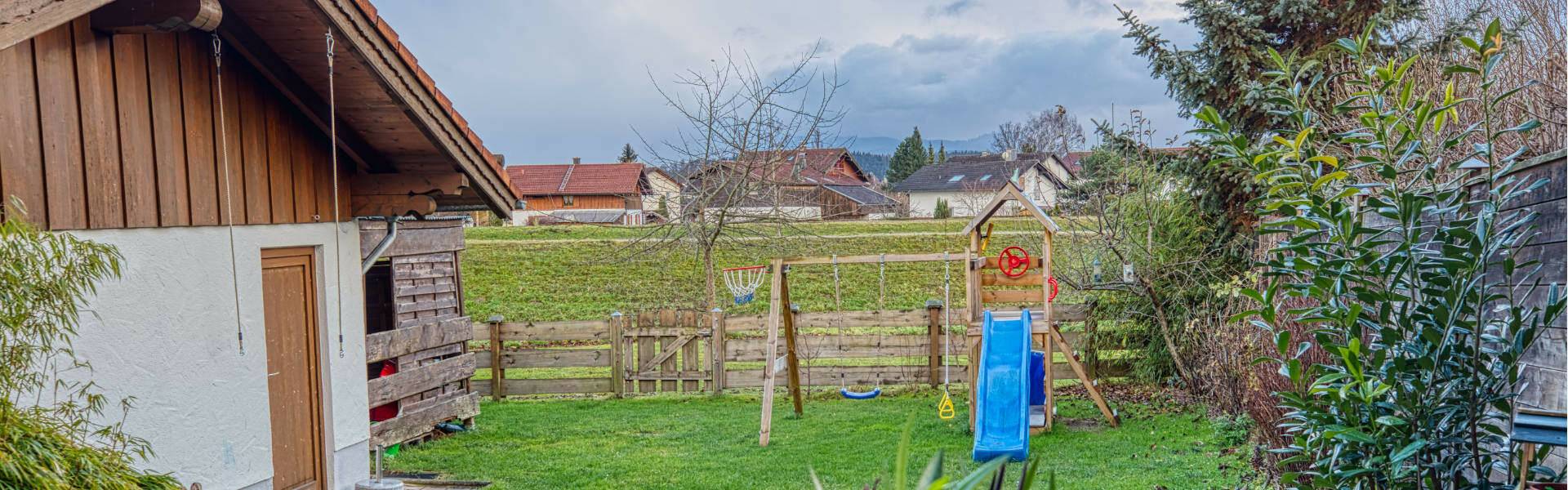 Wide panoramic view over a garden with a wooden shed, fence, and neighboring houses, as well as a blue play equipment.