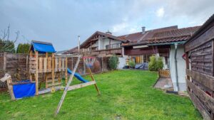Garden area with a wooden play tower including a slide and swing, surrounded by townhouses under a cloudy sky.
