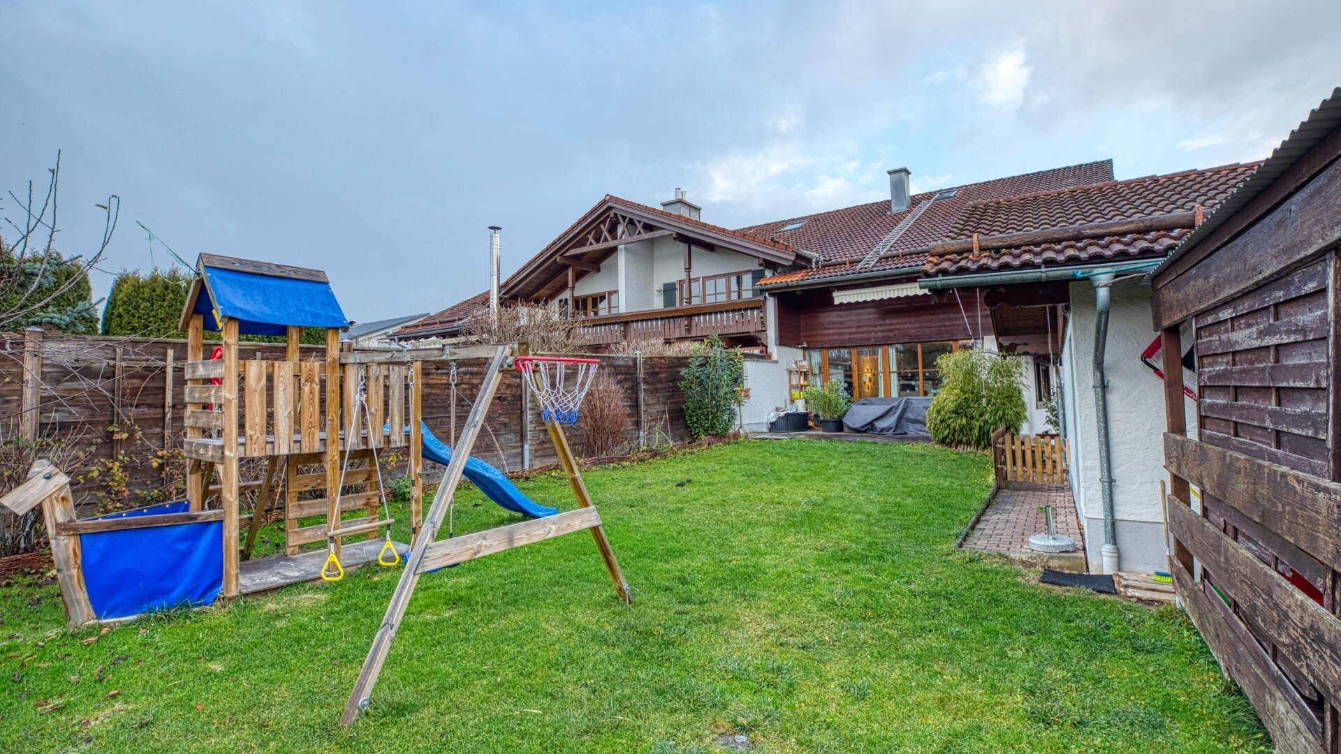 Garden area with a wooden play tower including a slide and swing, surrounded by townhouses under a cloudy sky.