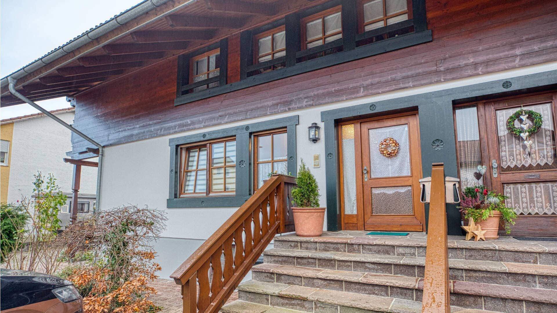 Exterior view of a house with wooden siding, a wooden entrance staircase, and decorative door wreaths in Bruckmühl.