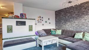 Bewohntes Wohnzimmer mit dunkler Eckcouch, Natursteinwand, weißem Kachelofen und Familienfotos an der Wand.