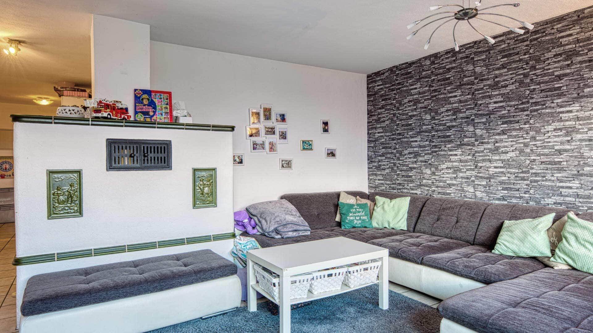 Lived-in living room with a dark corner sofa, natural stone wall, white tiled stove, and family photos on the wall.