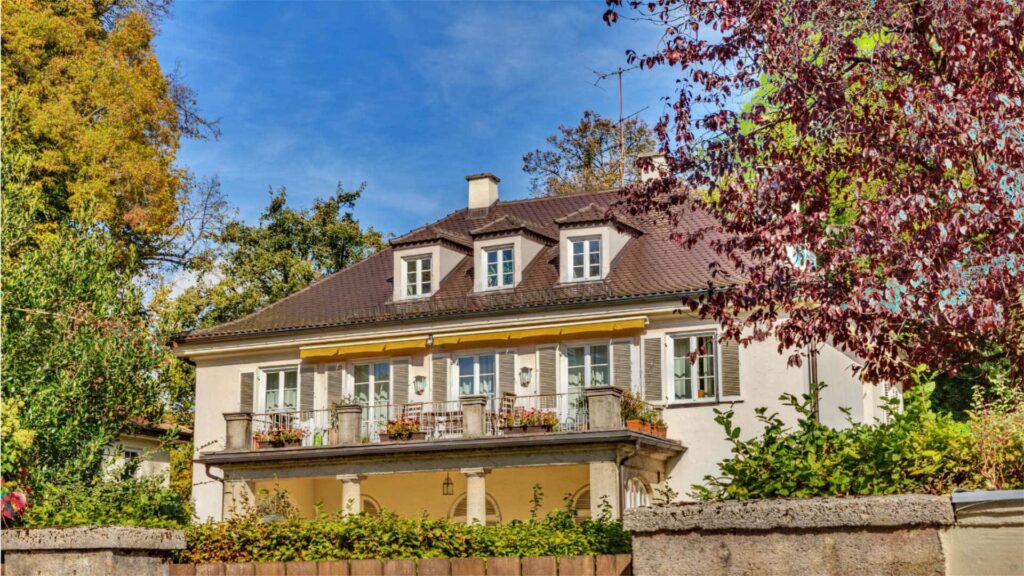 Cremefarbene Villa mit Balkon und Fensterläden, umgeben von herbstlich gefärbten Bäumen – Symbolbild zu Eigenheimversicherungen