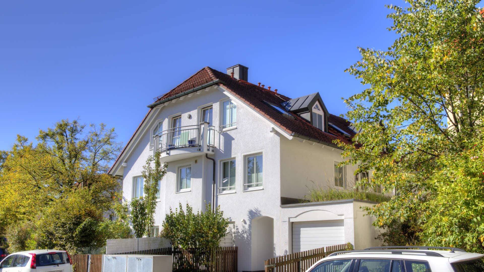 Weißes Haus mit rotem Ziegeldach, Bäumen und parkenden Autos an sonnigem Tag – Symbolbild zu Fehlern beim Immobilienverkauf