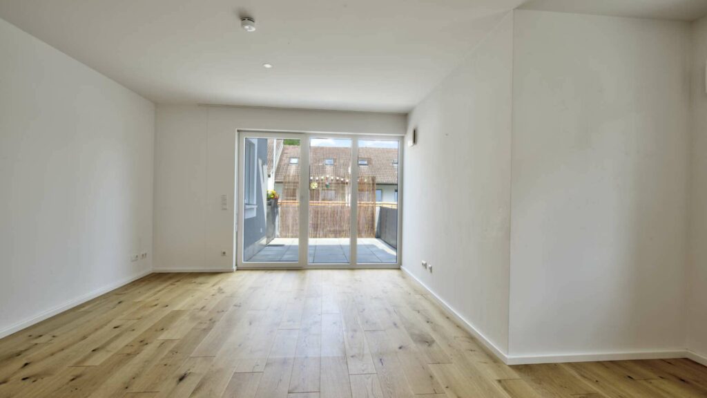 Leeres Wohnzimmer mit hellem Holzboden und Glastür zum Balkon in einer Eigentumswohnung in Gröbenzell, vermittelt von Rogers Immobilien