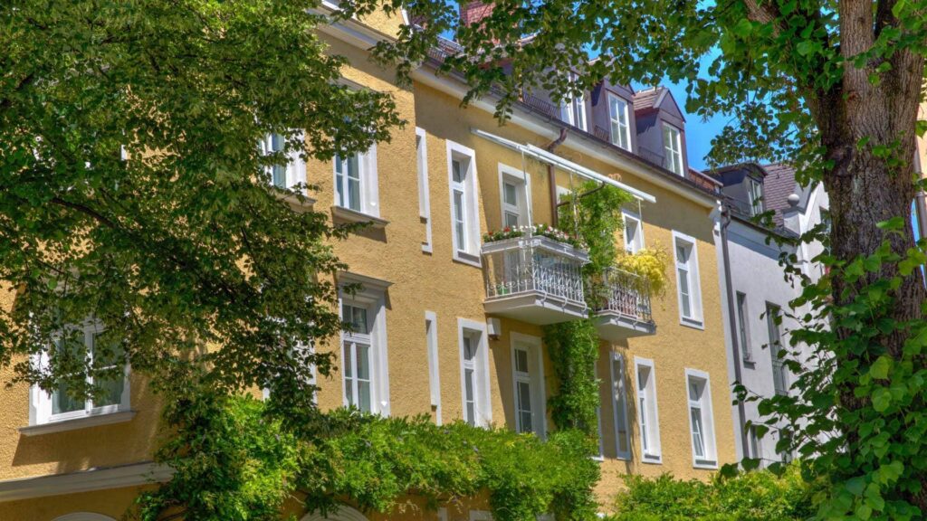 Ockerfarbenes Altbau-Wohnhaus mit weißen Fensterrahmen und kleinen Balkonen hinter Bäumen – Symbolbild zur Verkäuferhaftung