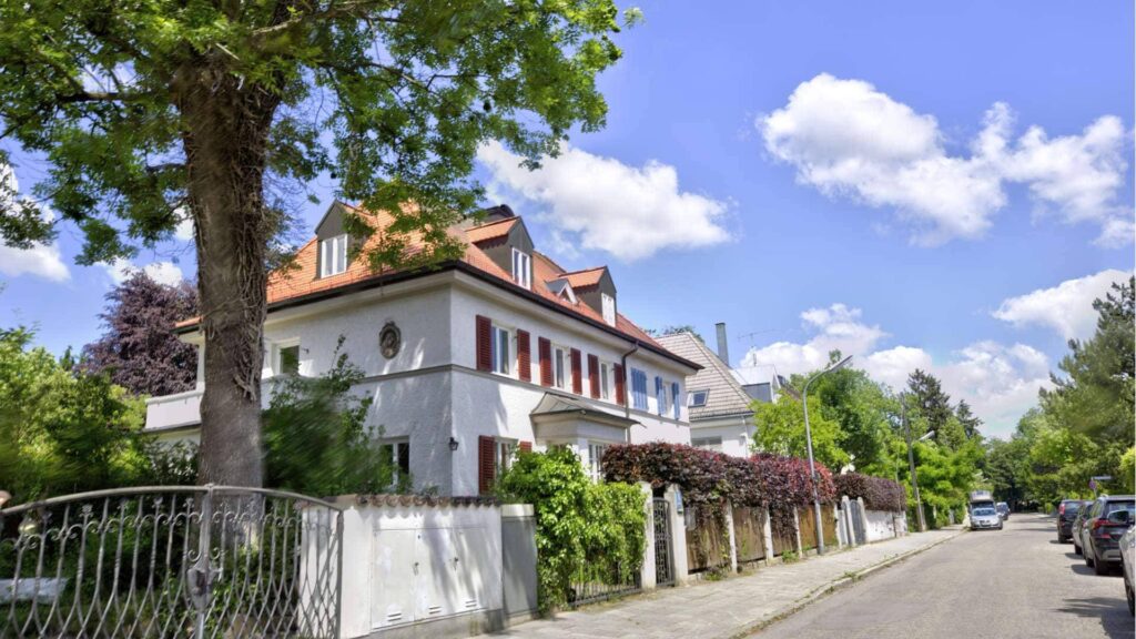 Weiße Villa mit orangefarbenem Dach, altem Baum und schmiedeeisernem Zaun an einer Straße – Symbolbild zum Außenbereich beim Hausverkauf