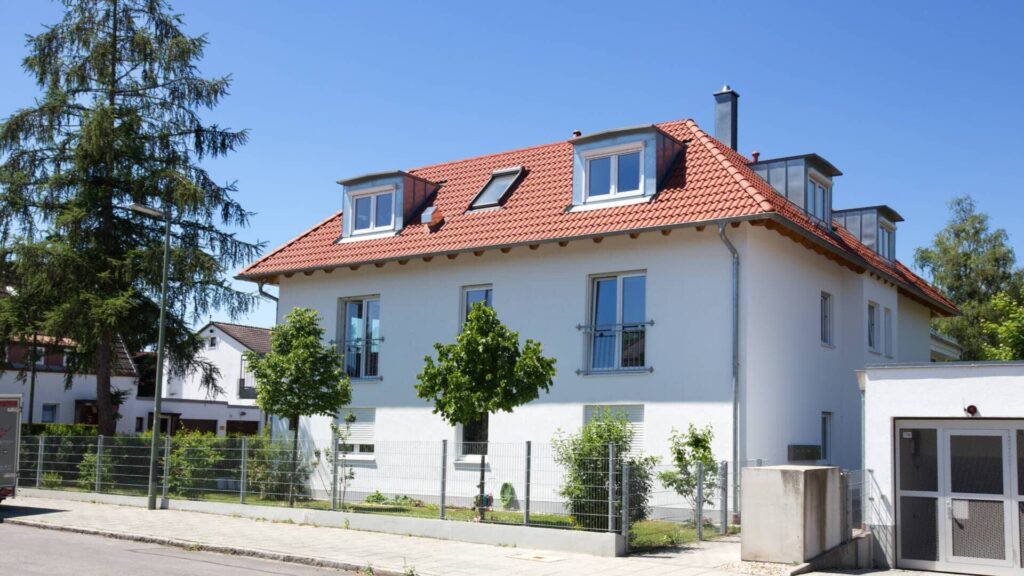 Weißes Wohnhaus mit rotem Ziegeldach, Dachgauben und Nadelbaum – Symbolbild zum Heizkosten sparen