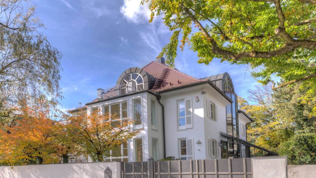 Cremefarbene Villa mit rundem Erker, weißem Zaun und buntem Herbstlaub – Symbolbild zum Heizöl sparen