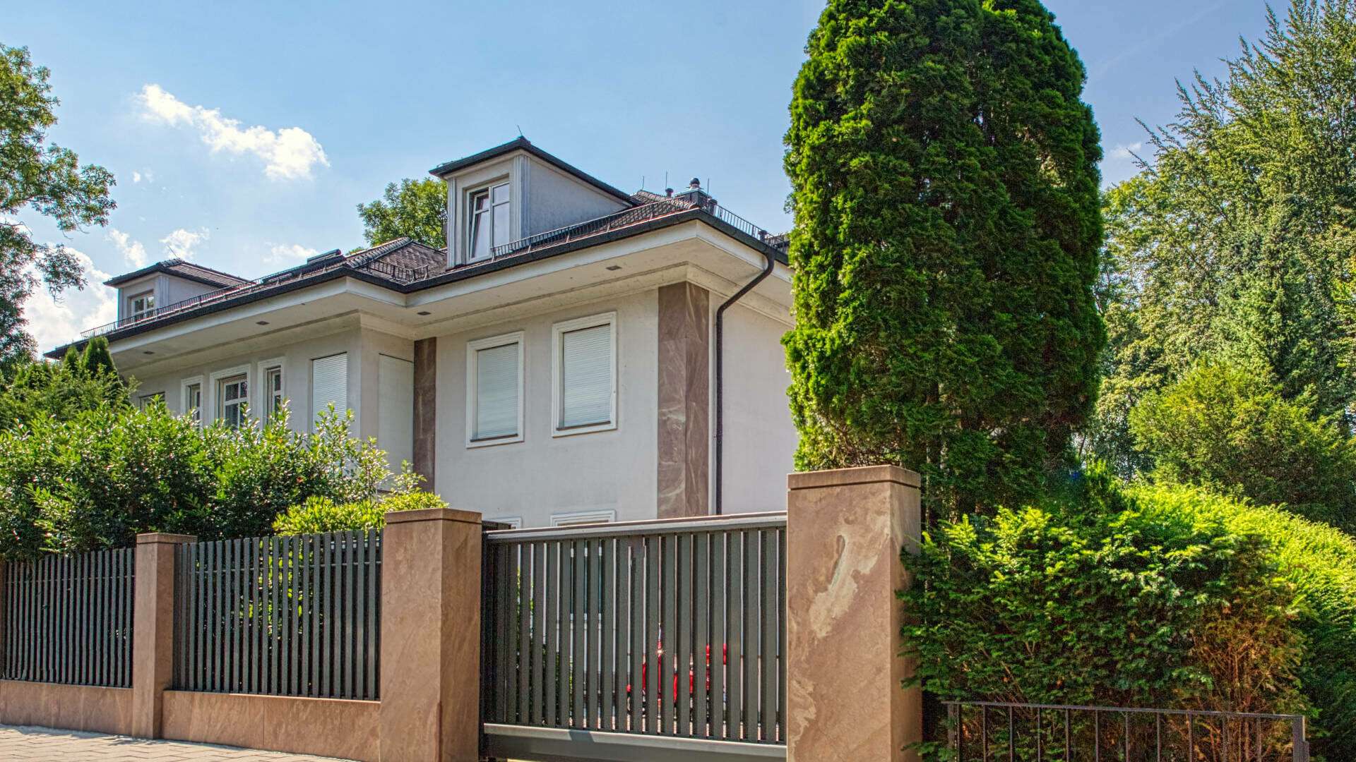Weiße Villa mit Dachgaube hinter schmiedeeisernem Zaun und Bäumen