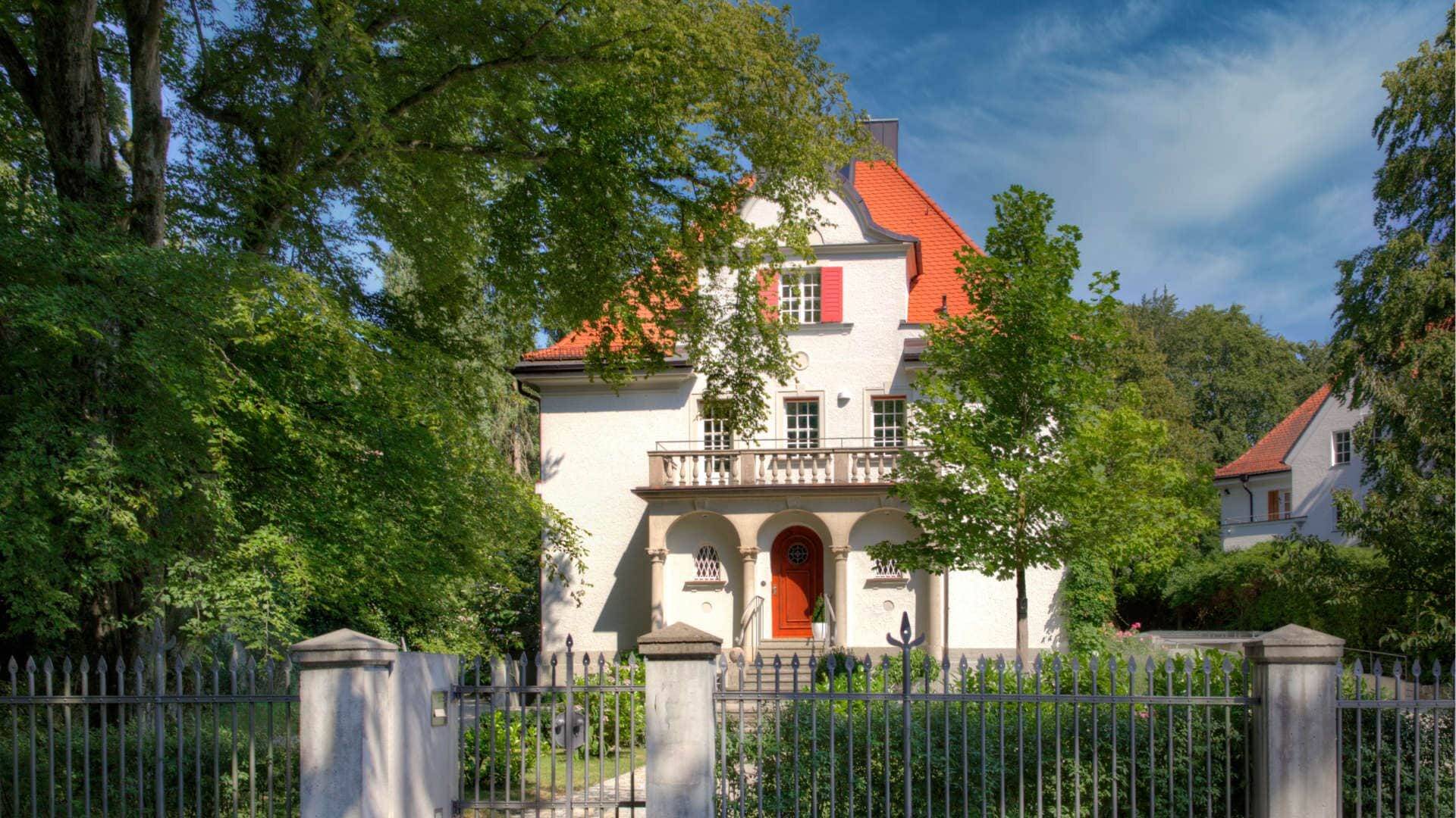 Weißes Haus mit rotem Dach, schmiedeeisernem Zauntor und rotem Eingang – Beispielbild zum Immobilienkaufvertrag
