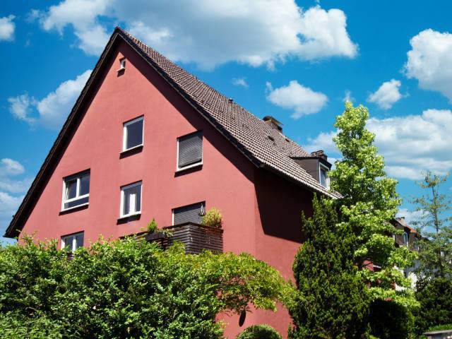 Rot verputztes Einfamilienhaus mit spitzem Giebel hinter grüner Hecke – Symbolbild zum Immobilienmarkt in Allach