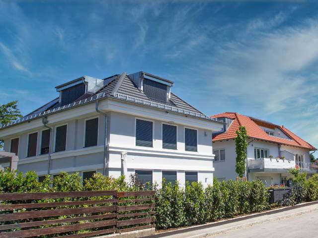Weißes Mehrfamilienhaus mit Gaube neben rotem Villendach hinter Hecke – Symbolbild zu Immobilienpreisen in Laim