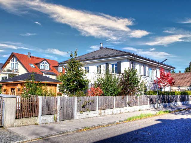 Weißes Wohnhaus mit Walmdach hinter Zaun neben rotem Nachbarhaus – Symbolbild zu Immobilienpreisen in Planegg