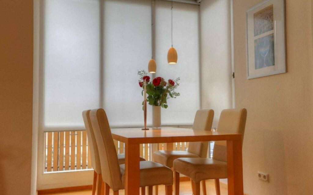 Gedeckter Glastisch mit Blumenstrauß und hellen Stühlen vor Fenster mit Rollo in einem Essbereich in Lerchenau.