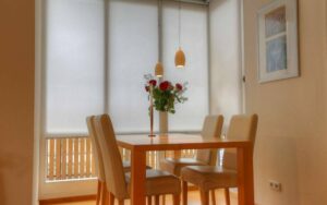 Gedeckter Glastisch mit Blumenstrauß und hellen Stühlen vor Fenster mit Rollo in einem Essbereich in Lerchenau.